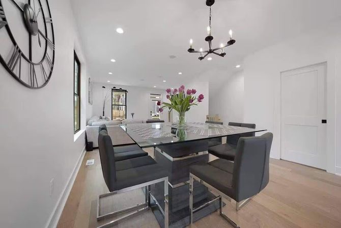 Bright modern open-plan dining room with a glass-top table, black leather chairs, vase of pink tulips, white walls, hardwood floors, wall clock and chandelier.