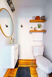 Compact modern bathroom with round gold-framed mirror, white floating vanity and toilet, wooden floating shelves holding potted plants and candles, a hanging white towel and warm hardwood floor.
