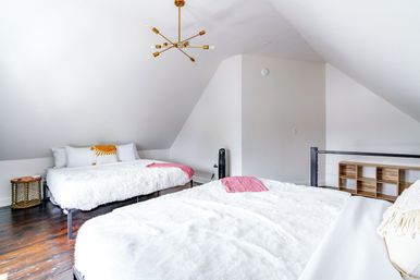 Cozy bright attic-style loft bedroom with two low-profile beds topped with fluffy white bedding and pink throws, sloped white ceilings, dark hardwood floors, and a brass modern chandelier.