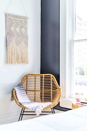 Cozy sunlit boho bedroom corner with a rattan chair draped in a striped throw, macramé wall hanging, black accent trim and a bright window.