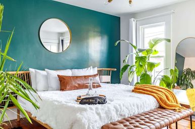 Sunlit modern bedroom with teal accent wall and round gold mirror, king bed layered with white faux-fur duvet, brown leather lumbar pillow, tufted leather bench, mustard throw, and lush indoor plants by a bright window.
