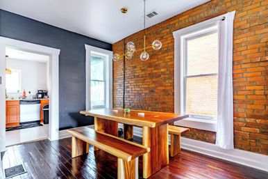 Bright urban dining area with exposed brick wall, live-edge wooden table and benches, globe pendant lights, tall windows with white curtains, and dark hardwood floors with a view into the kitchen.