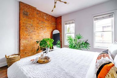 Sunny urban loft bedroom with exposed brick accent wall, white textured bedding, potted tropical plants, arched floor mirror, wicker basket and decorative tray