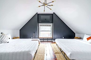 Bright modern attic bedroom with two white beds under sloped ceilings, a dark charcoal accent wall framing a central window bench, hardwood floors, woven rugs and a sputnik chandelier