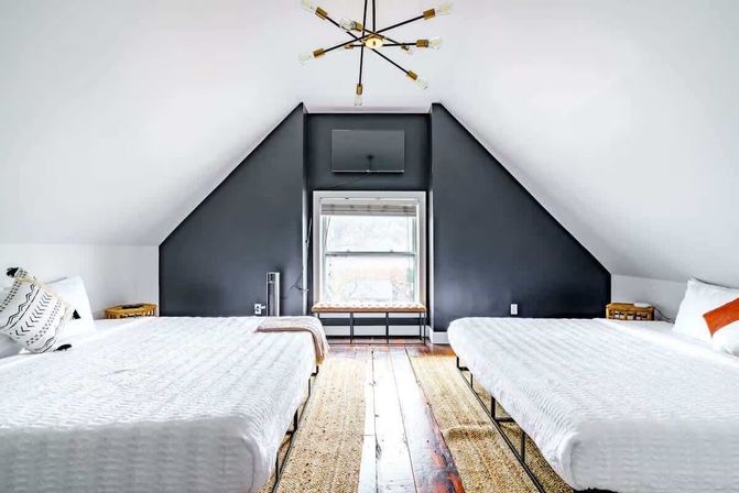 Bright modern attic bedroom with two white beds under sloped ceilings, a dark charcoal accent wall framing a central window bench, hardwood floors, woven rugs and a sputnik chandelier