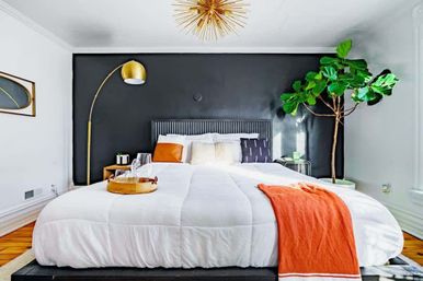 Modern bedroom with black accent wall, king bed dressed in white comforter and orange throw, gold arc floor lamp, sunburst chandelier, fiddle-leaf fig plant and wooden floors