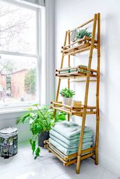 Sunlit bathroom corner with a bamboo ladder shelf by a large window — stacked mint towels, potted succulents and a leafy houseplant on the shelves, bright white tile floor and natural light