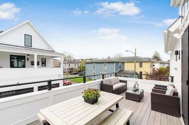 Sunny second-story rooftop deck with white picnic table and potted flowers, wicker lounge seating, glass railing and lanterns overlooking a quiet residential neighborhood under a blue sky