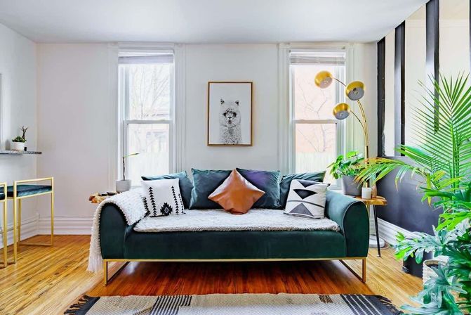 Bright, stylish urban living room with a green velvet sofa, patterned throw pillows, hardwood floors, gold arc floor lamp, potted plants, and a framed animal print between two windows.