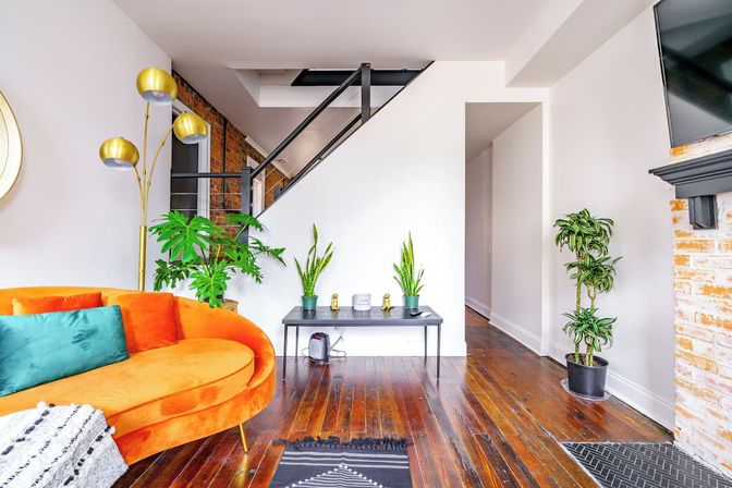 Urban loft living room with curved orange velvet sofa and teal cushion, gold arc floor lamp, exposed brick wall, black metal staircase, hardwood floors and potted indoor plants.