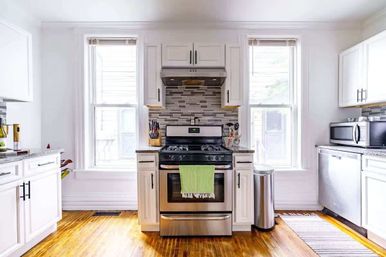 Sunny modern white kitchen with stainless steel gas range and green towel, mosaic tile backsplash, two tall windows, hardwood floors, microwave and dishwasher