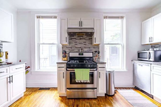Sunny modern white kitchen with stainless steel gas range and green towel, mosaic tile backsplash, two tall windows, hardwood floors, microwave and dishwasher