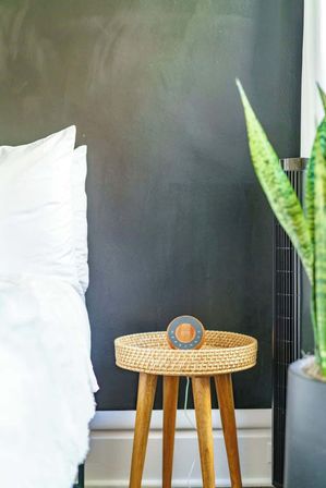 Cozy modern bedroom corner with white bedding against a dark accent wall, a woven round wooden bedside table holding a small orange smart speaker, and a tall snake plant in a gray pot.