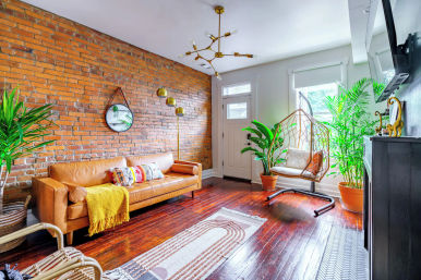 Cozy sunlit urban loft living room with exposed brick wall, tan leather sofa, hanging rattan chair, large potted plants and glossy hardwood floors