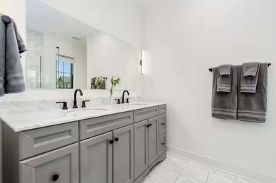 Modern bright bathroom interior with double-sink marble countertop, gray shaker vanity and black faucets, large wall mirror, vase of white flowers, and neatly folded gray towels on a wall bar.