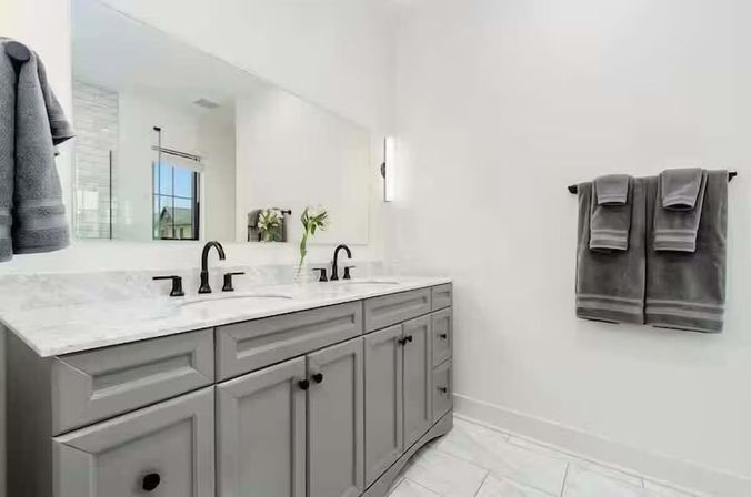 Bright modern master bathroom with double-sink gray vanity, marble countertop, matte black faucets, large mirror and neatly folded gray towels on a white wall.