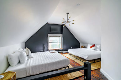 Cozy attic loft bedroom with two white-dressed beds, angled white ceiling and black accent wall with window and wall-mounted TV, mid-century chandelier, hardwood floors and woven rugs — bright, modern rustic vibe.