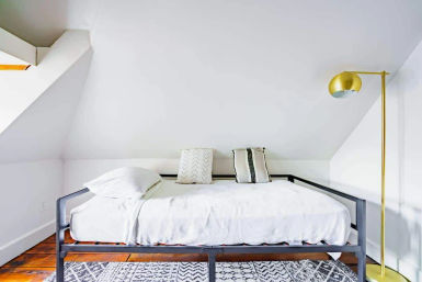 Cozy minimalist attic loft bedroom with a white daybed on a black metal frame, patterned throw pillows, a geometric rug, and a gold floor lamp under a sloped ceiling.