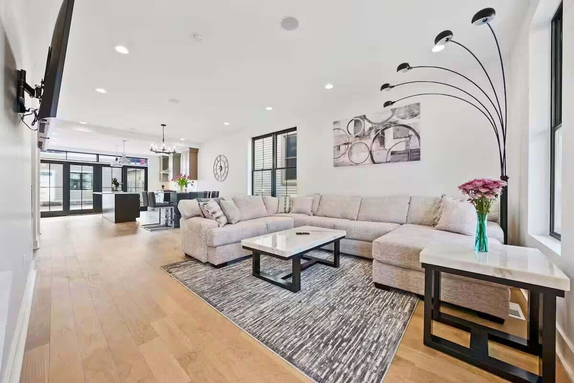 Bright modern open-concept living room and kitchen with light hardwood floors, L-shaped beige sectional, marble-top coffee table on a textured gray rug, multi-arm arc floor lamp, dining area and island in the background, and large windows flooding the space with natural light.