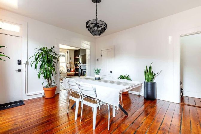Bright sunlit dining room with warm hardwood floors, white table topped with a fringed runner and four chairs, black woven chandelier, large potted plants, and an open view into a cozy living area with a hanging chair.