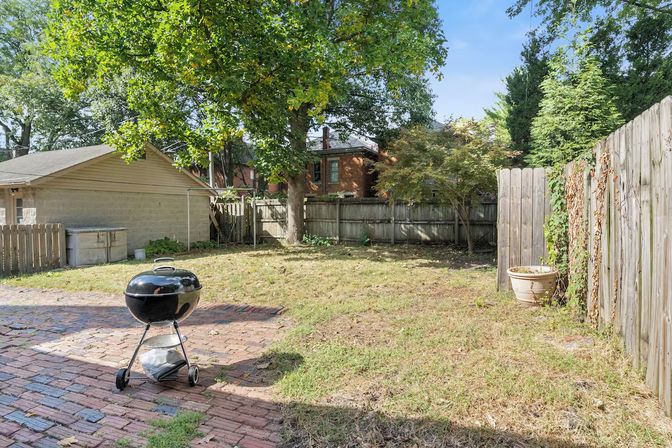 Sunny suburban residential backyard with brick patio and black kettle charcoal grill, grassy yard bordered by a wooden fence, large shade tree, potted plant, and detached garage.