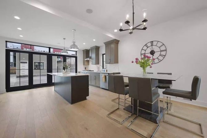 Open-concept modern kitchen with a central white island, gray cabinets, light hardwood floors, black-framed sliding glass doors, industrial pendant lights, dining bar with gray leather chairs and a wall clock with fresh tulips.
