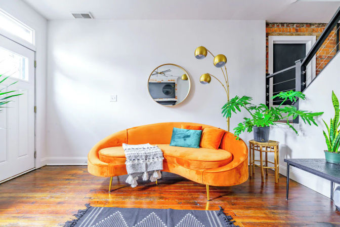 Bright contemporary loft living room with a curved orange velvet sofa on hardwood floors, teal throw pillow and knit blanket, round wall mirror, gold arc floor lamp and potted green plants against white walls.