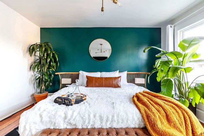 Cozy modern bedroom with teal accent wall and round mirror, wooden slatted headboard, fluffy white bedding, caramel leather pillow, mustard orange throw, tray with glassware, and large sunlit houseplants by the window.