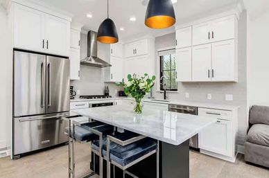 Bright modern white kitchen with a marble island, stainless steel French-door refrigerator, built-in range and dishwasher, black-and-gold pendant lights, velvet bar stools, and a vase of green-and-white flowers
