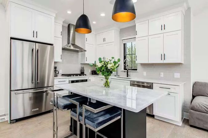 Bright modern white kitchen with a marble island, stainless steel French-door refrigerator, built-in range and dishwasher, black-and-gold pendant lights, velvet bar stools, and a vase of green-and-white flowers