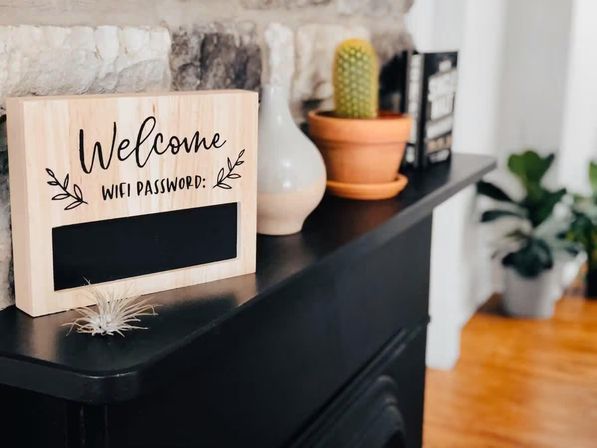 Wooden 'Welcome' Wi‑Fi password chalkboard on a black mantel with a small air plant, ceramic vase, terracotta potted cactus and books in a cozy living room