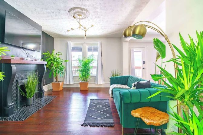 Sunny modern living room with an emerald velvet sectional, black fireplace and wall-mounted TV, brass arc floor lamp, hardwood floors, patterned runner rug and lush potted plants by bright windows.