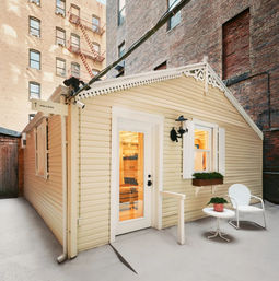 Cheerful yellow cottage tucked in an urban courtyard with a white door, window box, potted plant, patio chair, welcome sign and surrounding brick buildings with a fire escape