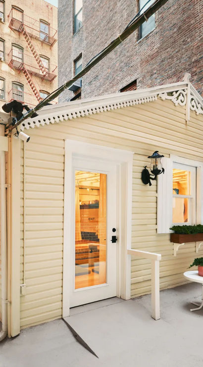 Cheerful yellow cottage tucked in an urban courtyard with a white door, window box, potted plant, patio chair, welcome sign and surrounding brick buildings with a fire escape