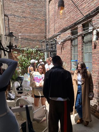 Sunlit urban brick alley patio with hanging string lights and potted plants, a small group of people in casual coats and sweaters chatting around chairs and a table.