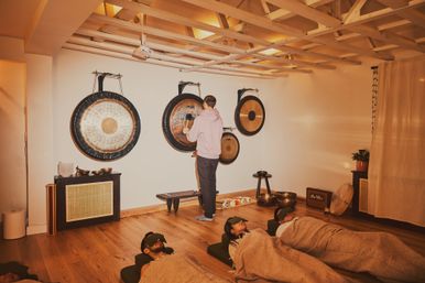 Participants recline with eye masks and blankets in a cozy wellness studio during a gong sound bath with hanging gongs and singing bowls on wooden floors.