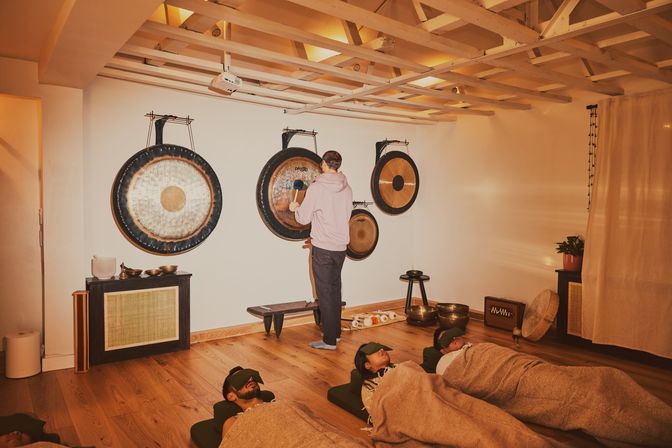 Participants recline with eye masks and blankets in a cozy wellness studio during a gong sound bath with hanging gongs and singing bowls on wooden floors.