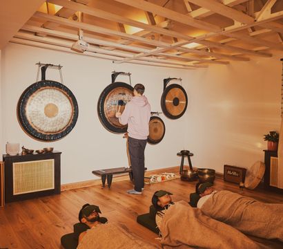 Participants recline with eye masks and blankets in a cozy wellness studio during a gong sound bath with hanging gongs and singing bowls on wooden floors.