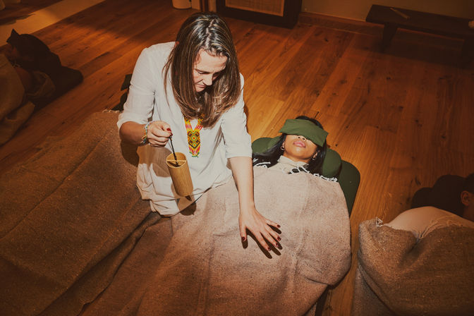 Practitioner performs a sound-healing session with a wooden sound instrument over a blindfolded client wrapped in a blanket in a cozy wellness studio with wooden floors.