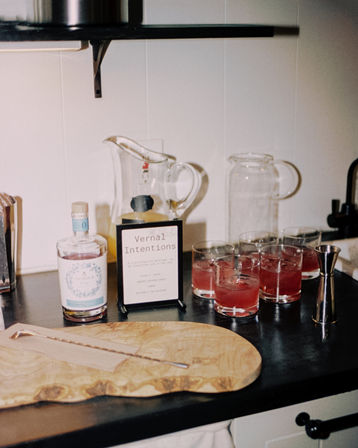 Home kitchen bar setup on a black countertop: wooden cutting board with a twisted bar spoon, clear botanical spirit bottle, sign reading "Vernal Intentions", glass pitchers, metal jigger and four pink spring cocktails in lowball glasses.