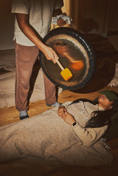 Cozy indoor wellness scene: person reclined on a hardwood floor under a blanket with an eye mask during a gong bath sound-healing session as a practitioner strikes a large gong with a mallet.