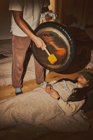 Cozy indoor wellness scene: person reclined on a hardwood floor under a blanket with an eye mask during a gong bath sound-healing session as a practitioner strikes a large gong with a mallet.
