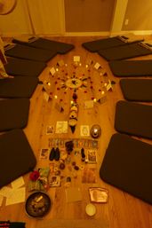 Warm amber-lit meditation circle on a hardwood floor with gray floor cushions arranged around a central mandala of candles, rose petals, oracle cards, crystals and small ritual objects for a group ceremony.