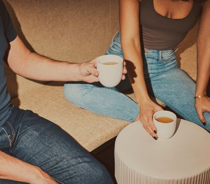 Two people in jeans sharing white ceramic mugs of tea on a beige sofa beside a round coffee table in a cozy living room.