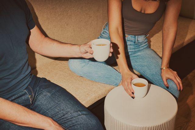 Two people in jeans sharing white ceramic mugs of tea on a beige sofa beside a round coffee table in a cozy living room.