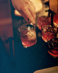 Hand using a jigger to pour amber craft cocktails over ice into short glasses on a dimly lit bar — moody nightlife scene