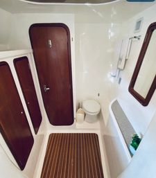 Cozy yacht bathroom (marine head) with white fiberglass walls, teak-striped floor mat, rounded mahogany cabin doors, compact toilet, wall mirror and small shelf with plant.