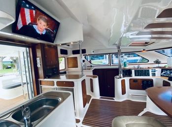 Sunlit luxury yacht interior with teak-style flooring, stainless galley sink, warm wood cabinetry, helm with navigation panels, large windows overlooking a marina, and a wall-mounted TV.