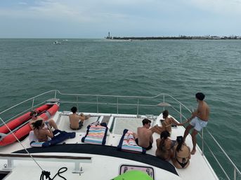 Group of people relaxing on the bow of a white yacht with striped beach towels and a red inflatable dinghy, looking out over teal coastal waters toward a distant island with a lighthouse under a cloudy sky — tropical boating vacation scene.