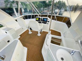 Flybridge cockpit of a white motor yacht with teak-style deck, twin captain chairs, helm with navigation screens, wet bar sink, and palm-tree marina in the background
